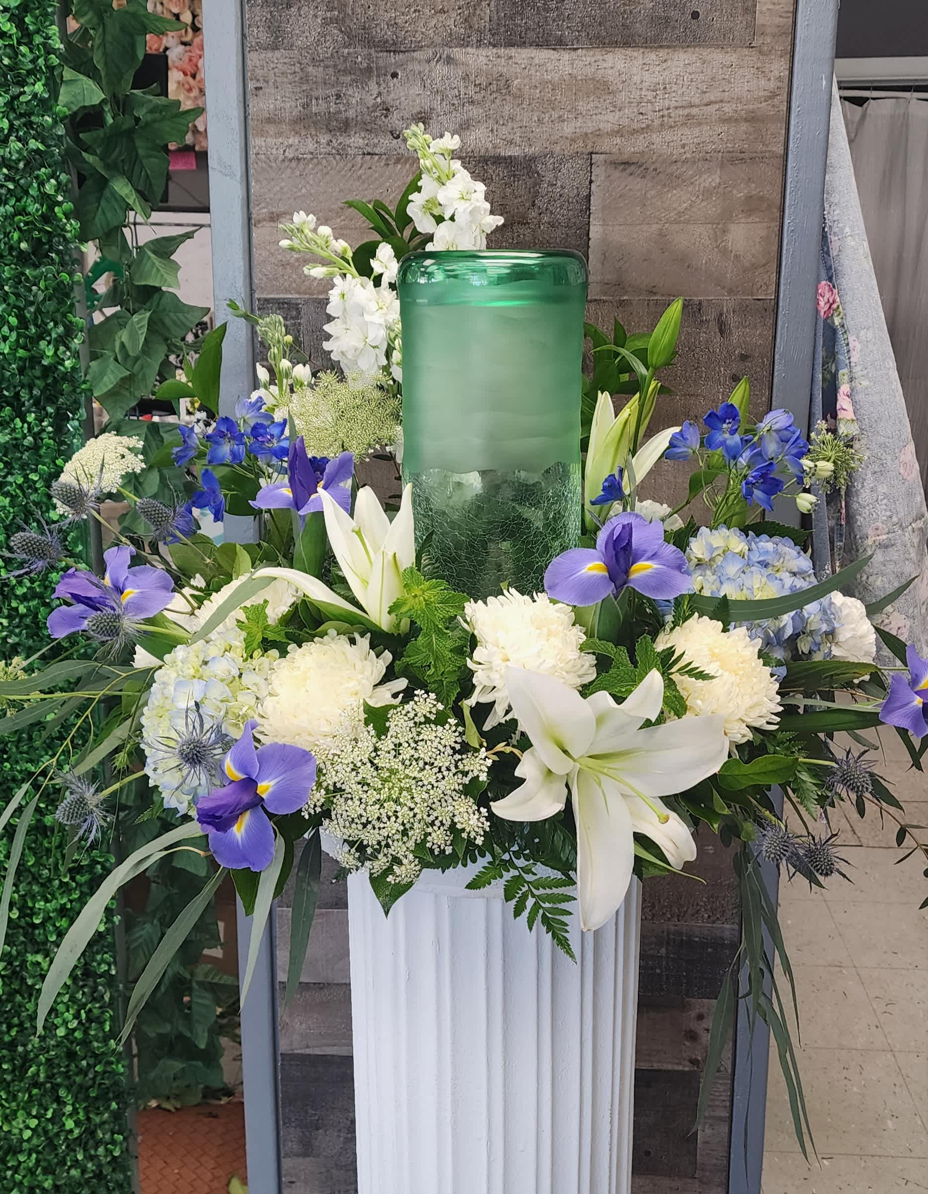 White lilies and blue-purple flowers arranged around a green glass candle holder