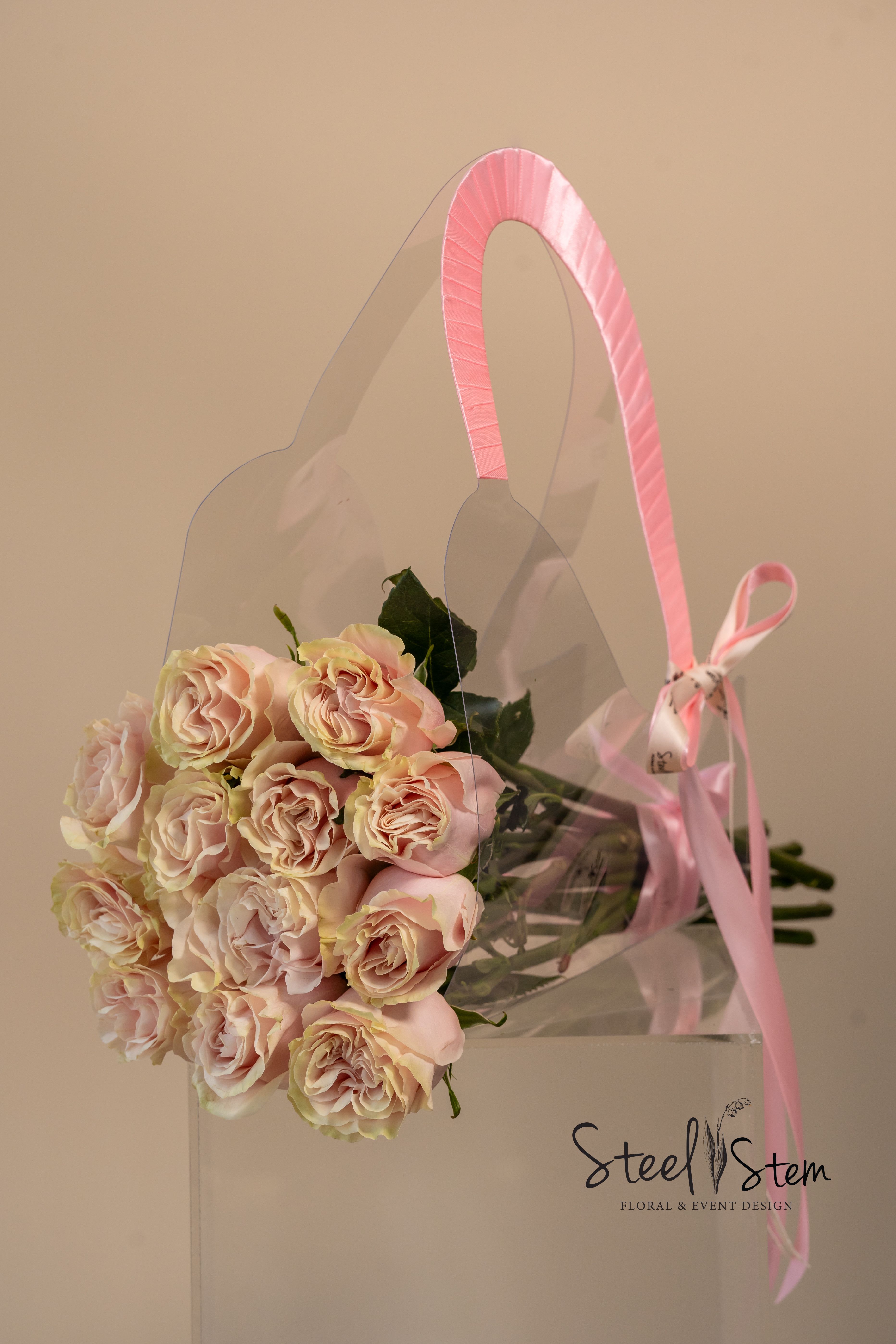 A bouquet of pale pink roses in a clear gift bag with a pink ribbon handle.