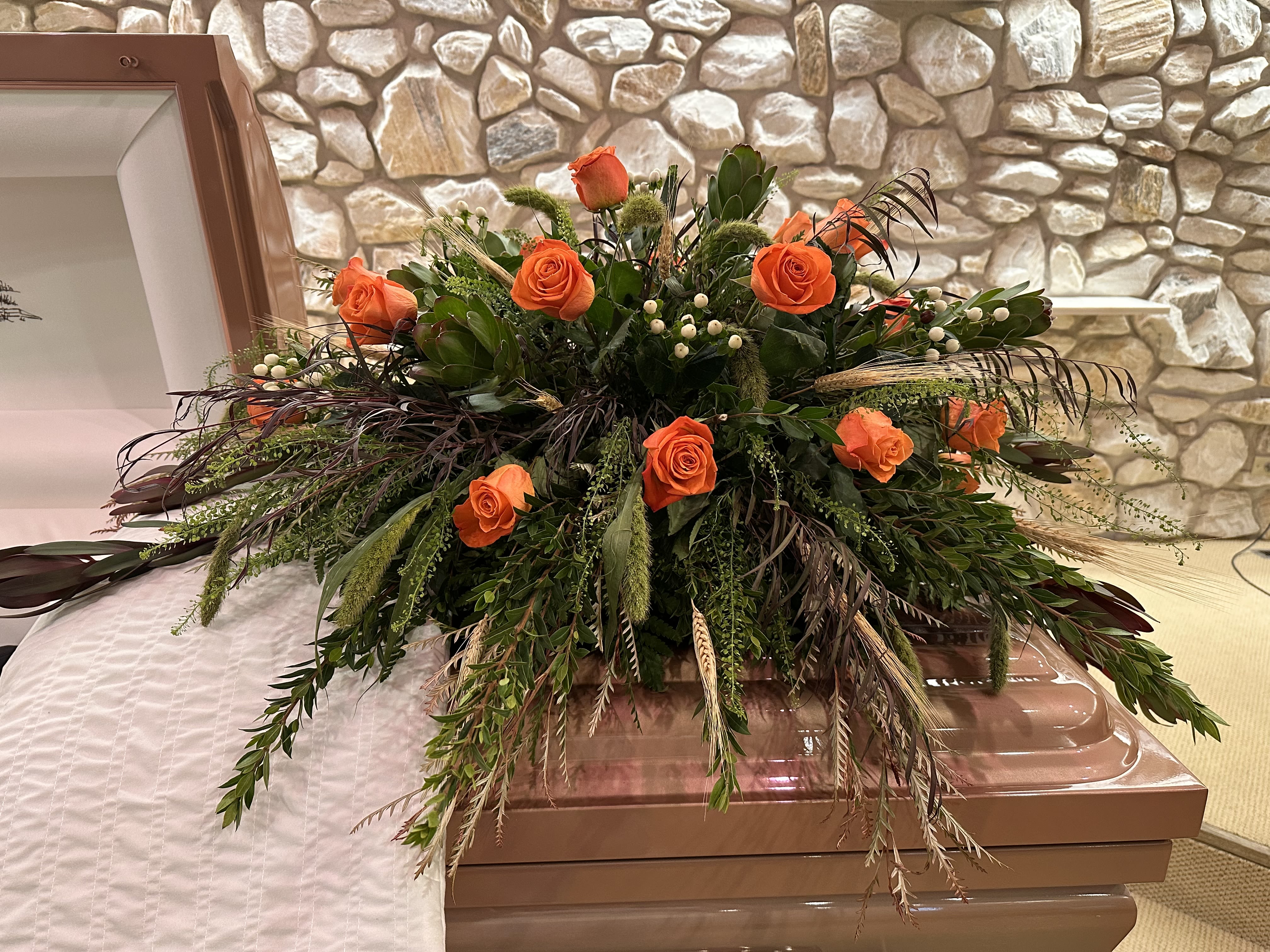 Everlasting Meadow - Bright clusters of orange roses form a warm, dignified casket spray designed for a funeral or memorial service. The arrangement reads as a solemn yet uplifting tribute-the vivid orange blooms draw the eye across the top of the casket while long, cascading greenery and trailing elements create a graceful, formal silhouette that honors the person you're remembering.  This casket spray pairs deep green foliage with textured filler and small white berry-like accents to add contrast and gentle sparkle against the roses. Purple-tinted touches and varied leaf shapes give depth and a refined color palette of orange, green, white, and purple. The design is broad and expansive, intended to spill slightly over the casket edges for visible presence during the service.  Choose this arrangement when you want the person you're thinking of to be remembered with warmth and dignity-an arrangement that feels both personal and respectful. Our team prepares and conditions each spray for longevity; please let us know the casket dimensions and service timing so we can ensure the piece is appropriately sized and delivered for the memorial.