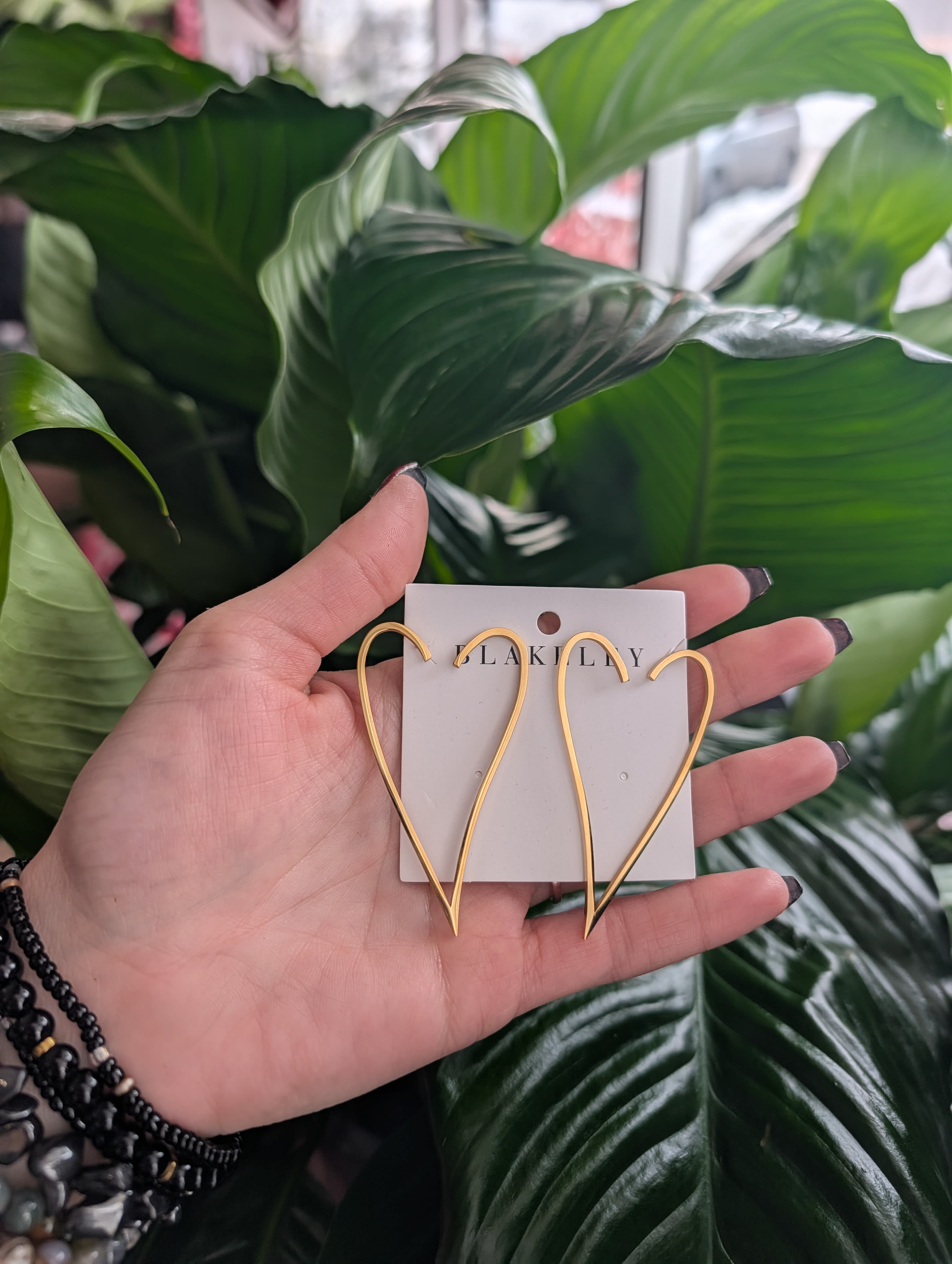 Hand holding gold heart-shaped earrings in front of large green leaves.