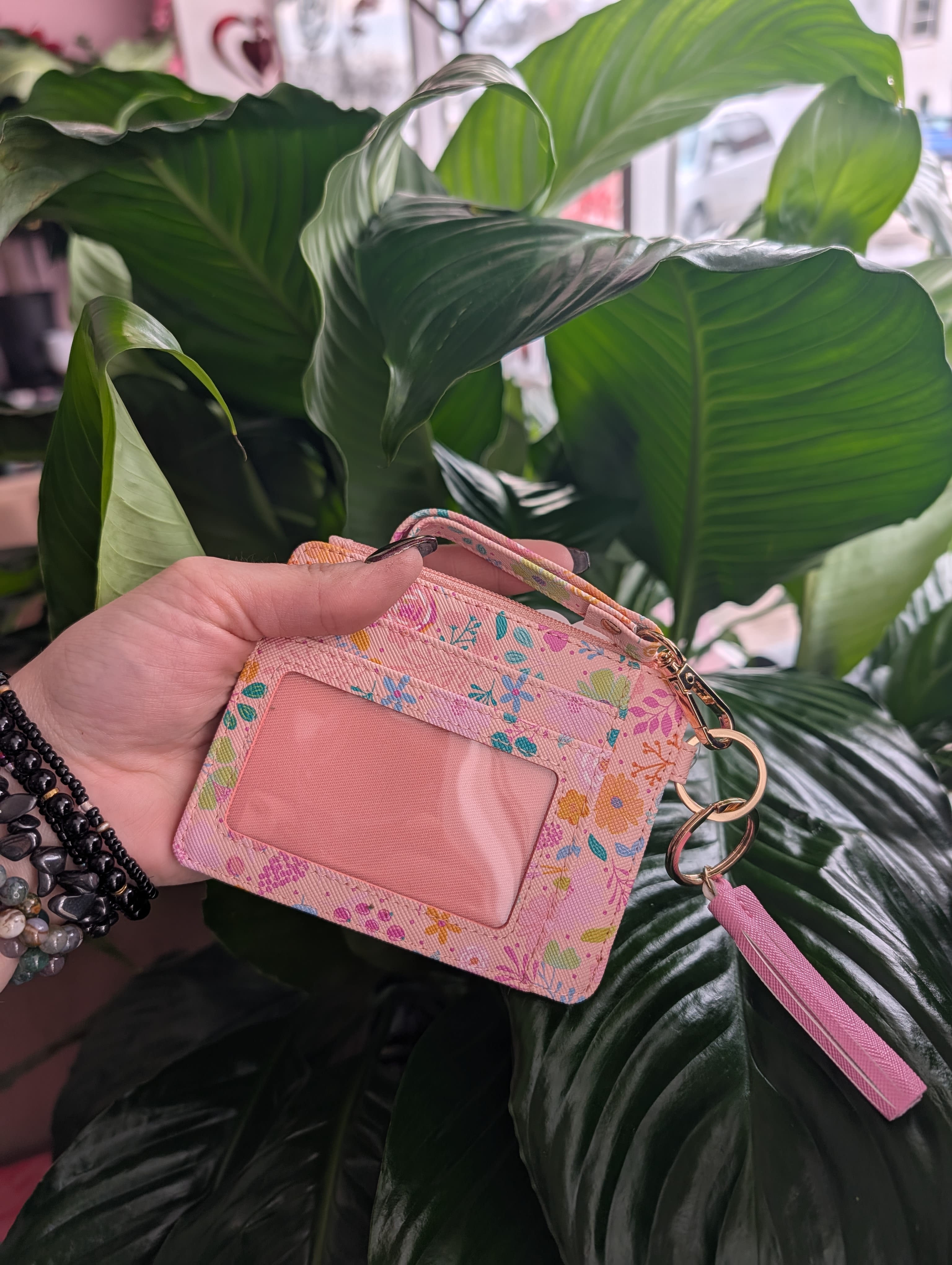 Hand holding a pink floral ID wallet in front of large green leaves.