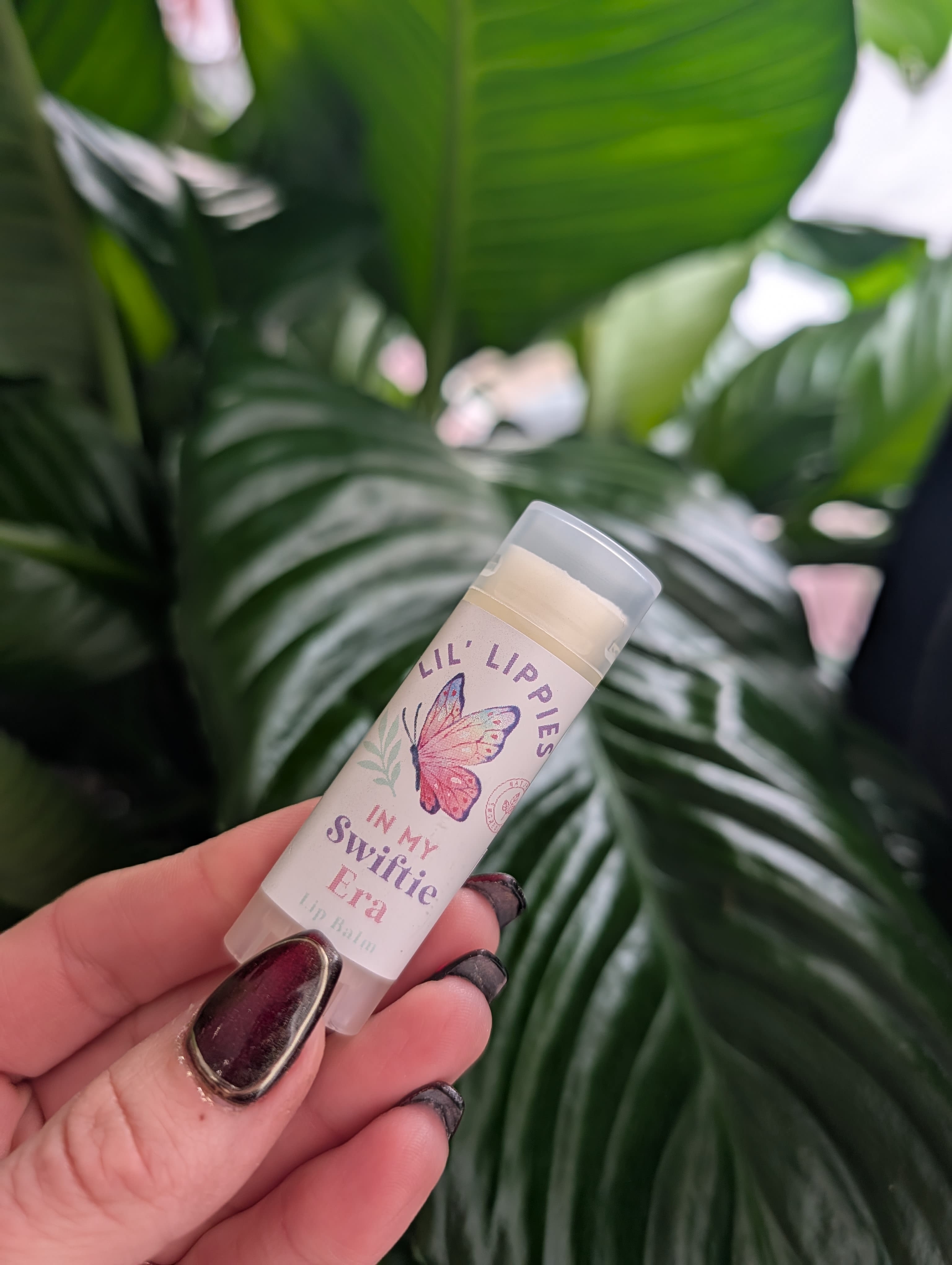 Hand holding a lip balm tube in front of large green leaves.
