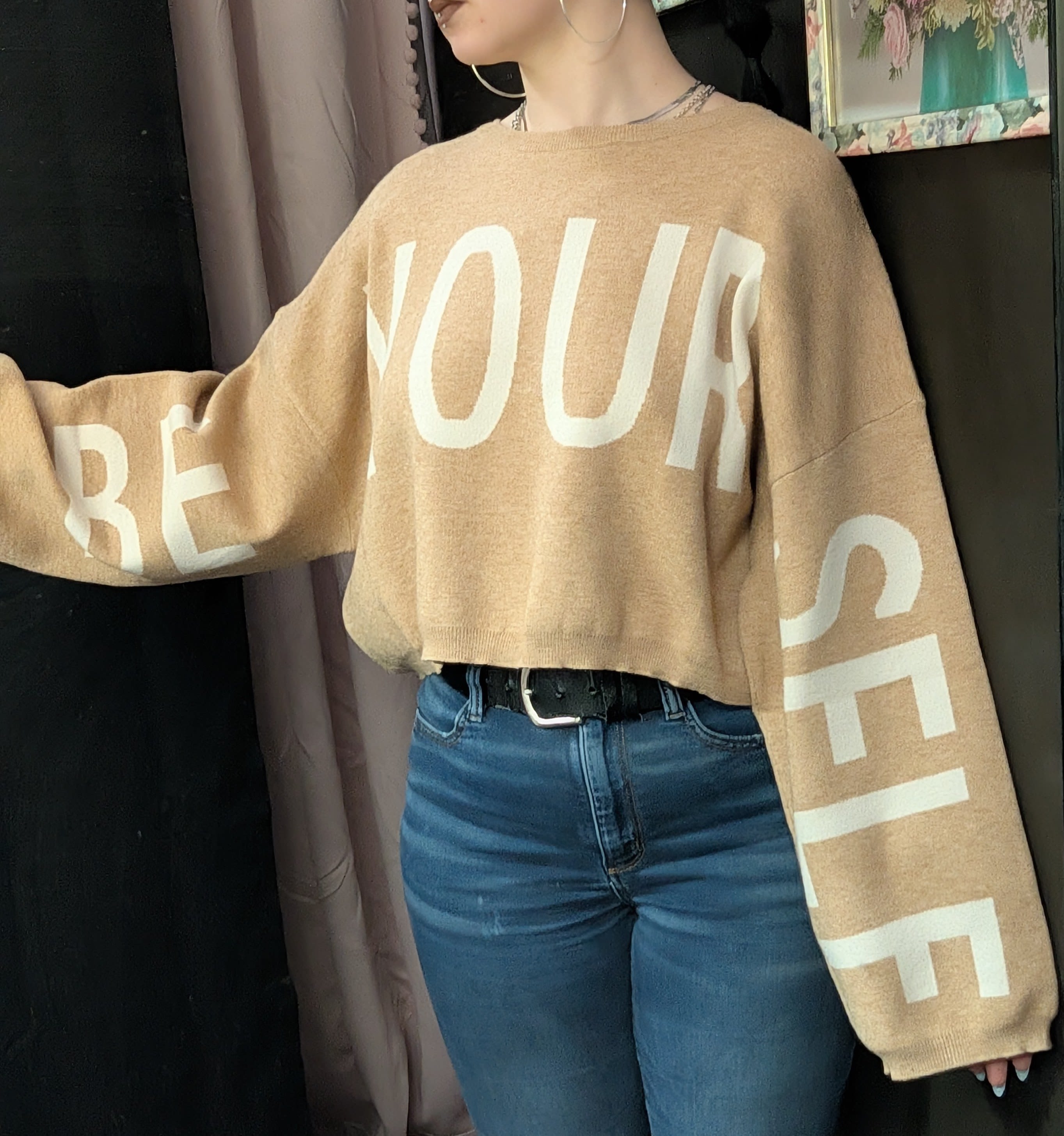 Person wearing a tan sweater with large white lettering and blue jeans