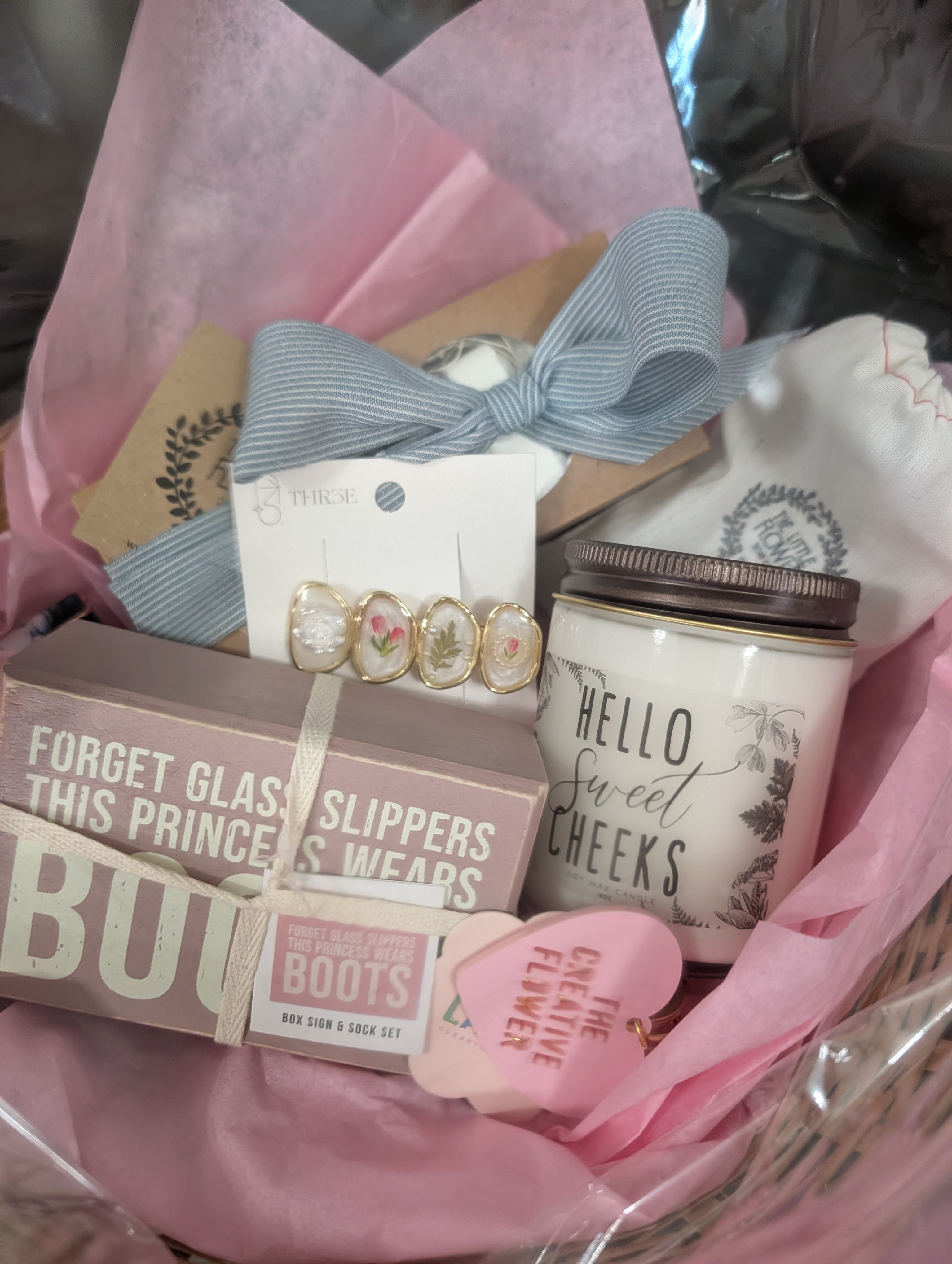 Pastel pink gift basket with candle, earrings, ribbon, and boxed sign set arranged in tissue paper.