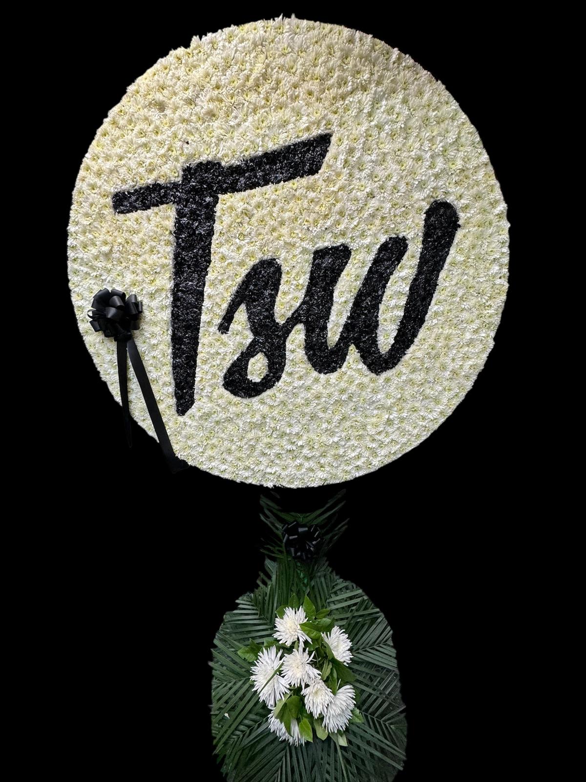 Round white floral tribute with black lettering and ribbon, mounted above a small floral base