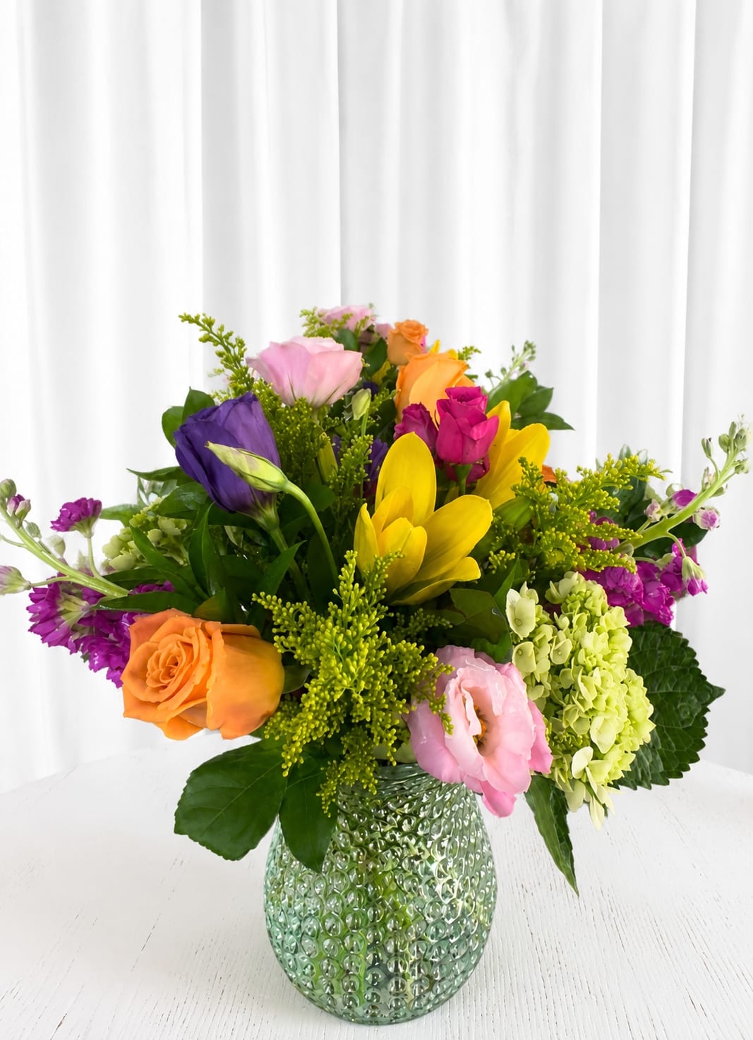 Rainbow  - A lively burst of color that feels like a garden in full cheer-this mixed-color flower arrangement is perfect as a birthday bouquet, a congratulations gift, or a bright spring surprise for someone special. The rounded, abundant composition immediately reads joyful and celebratory, the kind of bouquet that makes the person you're thinking of feel truly seen and celebrated.  Roses, tulips, lisianthus, hydrangeas and other seasonal blooms are gathered tight for a full, textured look in shades of pink, yellow, orange, purple and fresh green. Varied bloom sizes and soft to ruffled textures give the arrangement depth and movement, while the textured green glass vase grounds the design with a weighty, decorative finish that can be reused long after the flowers fade. The overall presentation is fresh, cheerful, and made to brighten a table, desk, or entryway.  Ideal for birthdays, new jobs, housewarmings, or just because, this arrangement sends a clear message of joy and celebration to the recipient. Delivered ready to display in its glass vase, enjoy simple care-fresh water, a cool spot away from direct sun and drafts-and replace water every other day to keep blooms looking their best.   If any design elements are of major importance to your order, please include them in the florist instructions at checkout or contact us to ensure availability.