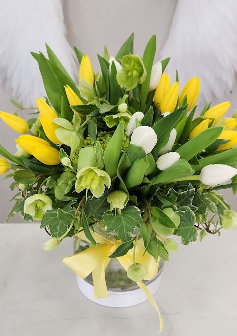 Yellow and white tulip arrangement with green blooms in a round vase with ivy and a yellow ribbon bow.