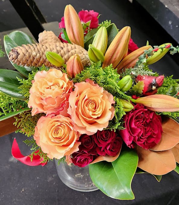 Peach and red roses with orange lilies, pinecones, and a red cardinal pick in a clear vase.