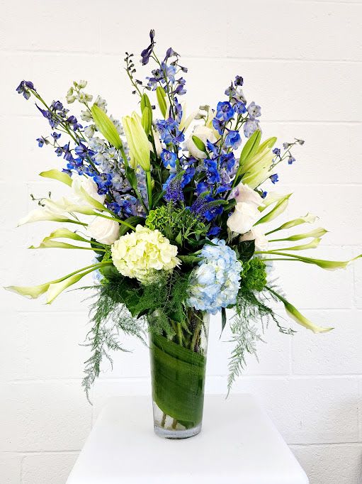 Tall blue and white flower arrangement with lilies, delphinium, and hydrangeas in a clear glass vase