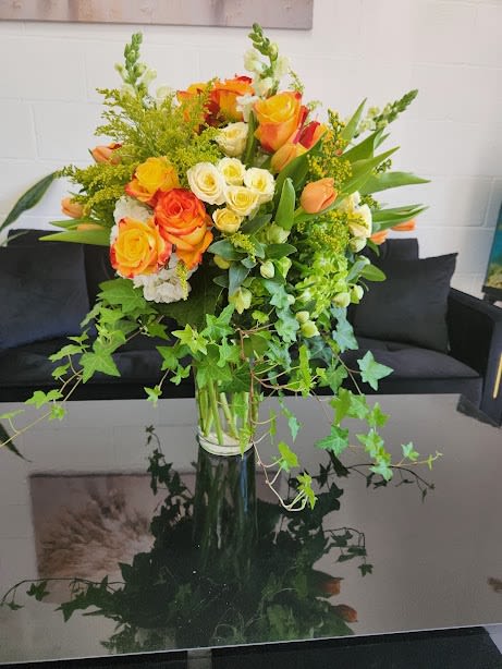 Tall vase of yellow and orange roses with white blooms and trailing ivy