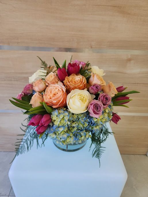 Short round arrangement of peach and cream roses, pink tulips, and blue hydrangeas in a clear glass vase