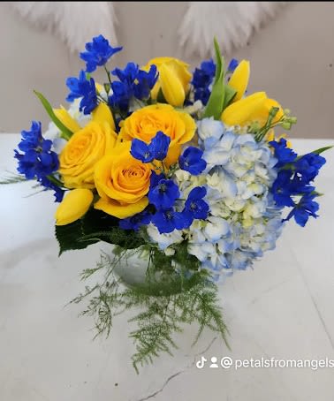 Low arrangement of yellow roses and tulips with blue delphinium and hydrangea in a clear glass vase