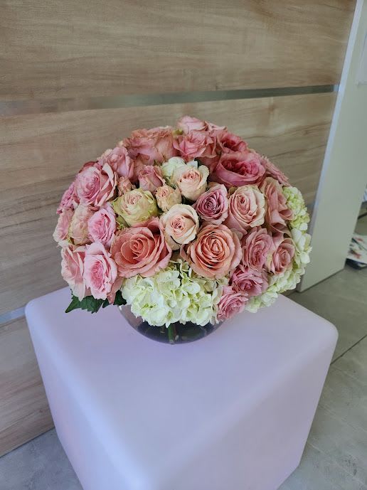 Round arrangement of pink roses and white hydrangeas in a clear glass bowl vase on a pale cube stand