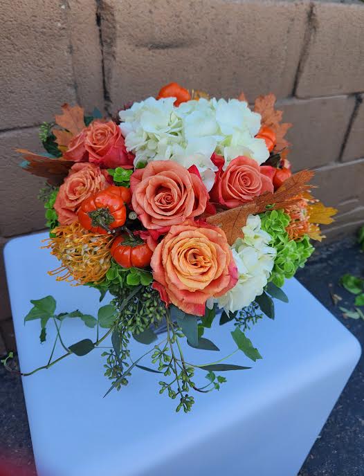 Low arrangement of orange roses, white hydrangeas, mini pumpkins, and trailing vines in a compact mound.