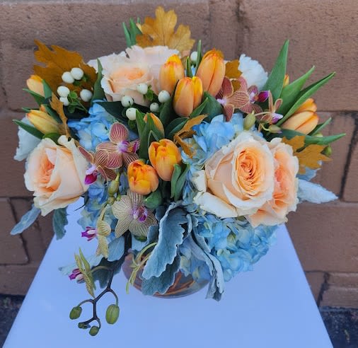 Low arrangement of peach roses, orange tulips, blue hydrangeas, and orchids in a clear glass vase