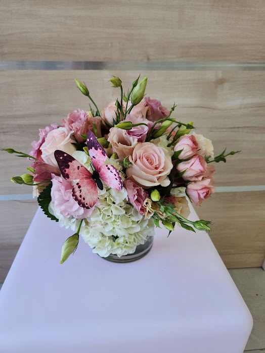 Low arrangement of blush pink roses and lisianthus with white hydrangeas in a glass vase and a pink butterfly accent