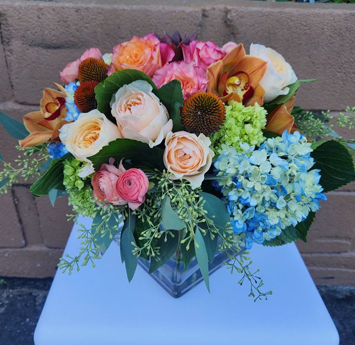 Low mixed arrangement of peach roses, blue hydrangeas and orchids in a square glass vase