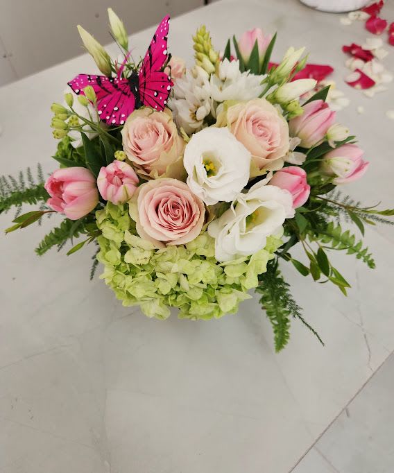 Round arrangement of blush roses, white blooms, pink tulips, and green hydrangeas with a pink butterfly pick