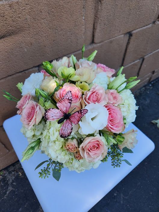 Round pastel arrangement of pink roses, white hydrangeas, and lisianthus with a pink butterfly decoration