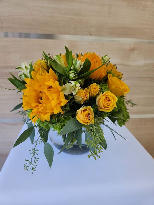 Low arrangement of yellow roses and gerbera daisies in a clear glass vase with green accents