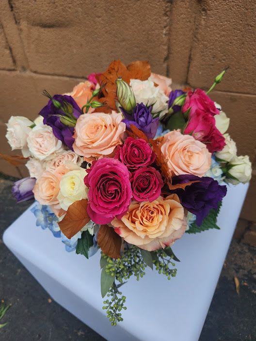 Round bouquet of pink and peach roses with purple lisianthus and blue accents on a white pedestal