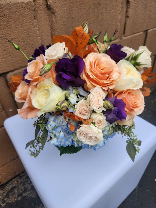 Compact arrangement of peach and cream roses, purple lisianthus, and blue hydrangea with orange leaves
