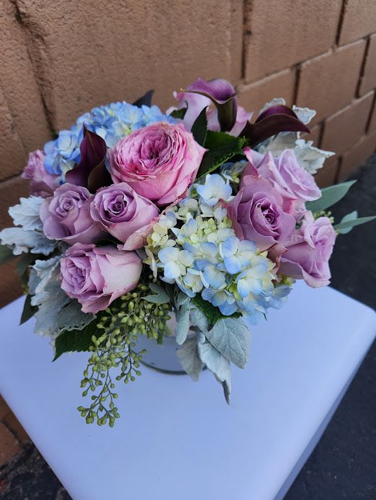 Low vase arrangement of pink and lavender roses, blue hydrangeas, and dark purple calla lilies.