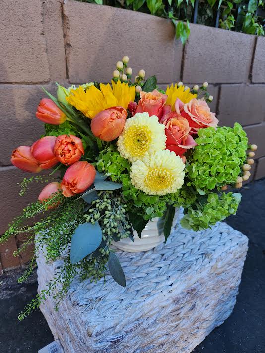 Low arrangement of orange tulips, yellow sunflowers, roses, mums and green hydrangea in a white vase