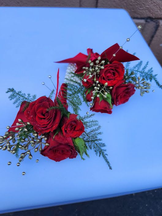 Two red rose wrist corsages with gold bead accents and ribbon on a light blue surface