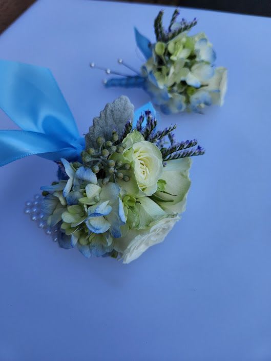 Blue and white wrist corsages with roses and hydrangea, accented with ribbon and pearls on a light background