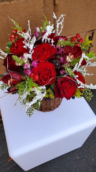 Low arrangement of red roses, orchids, and pink blooms with white frosted accents in a rustic bark container
