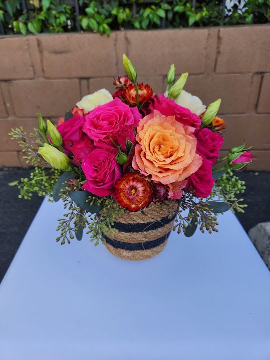 Compact arrangement of hot pink and peach roses in a striped woven basket container