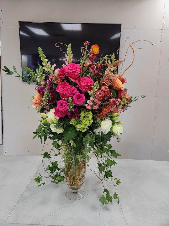 Tall arrangement of hot pink roses and mixed flowers in a clear glass vase with trailing ivy.