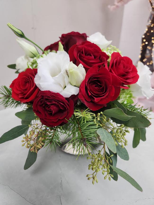 Low arrangement of red roses and white lisianthus with holiday greenery in a silver vase