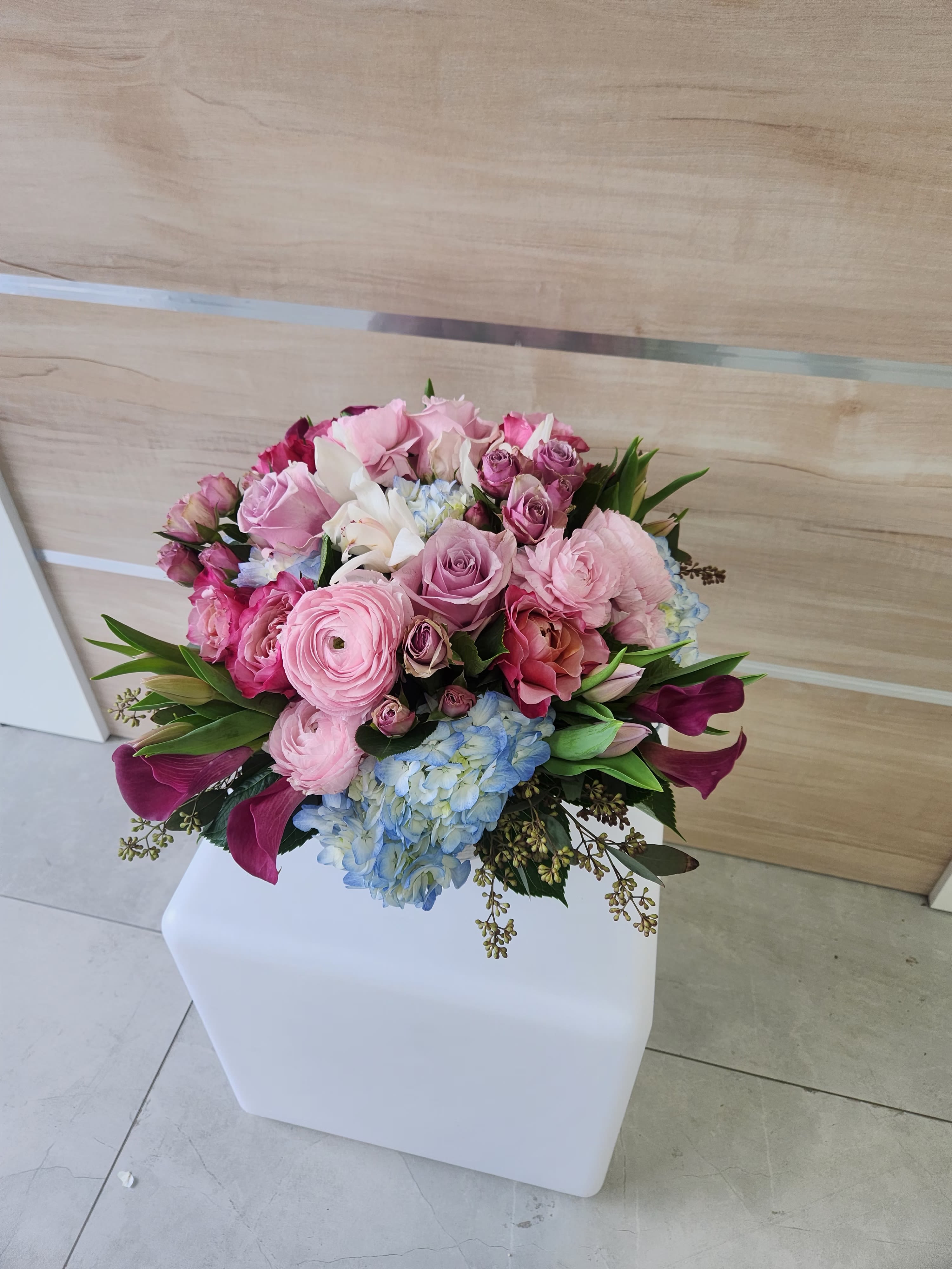 Rounded bouquet of pink roses, ranunculus, lilies and blue hydrangeas in a compact arrangement.