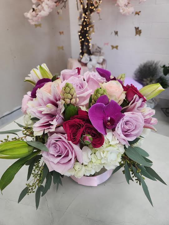 Low round arrangement of pink and purple roses, orchids, and white hydrangea in a hatbox container
