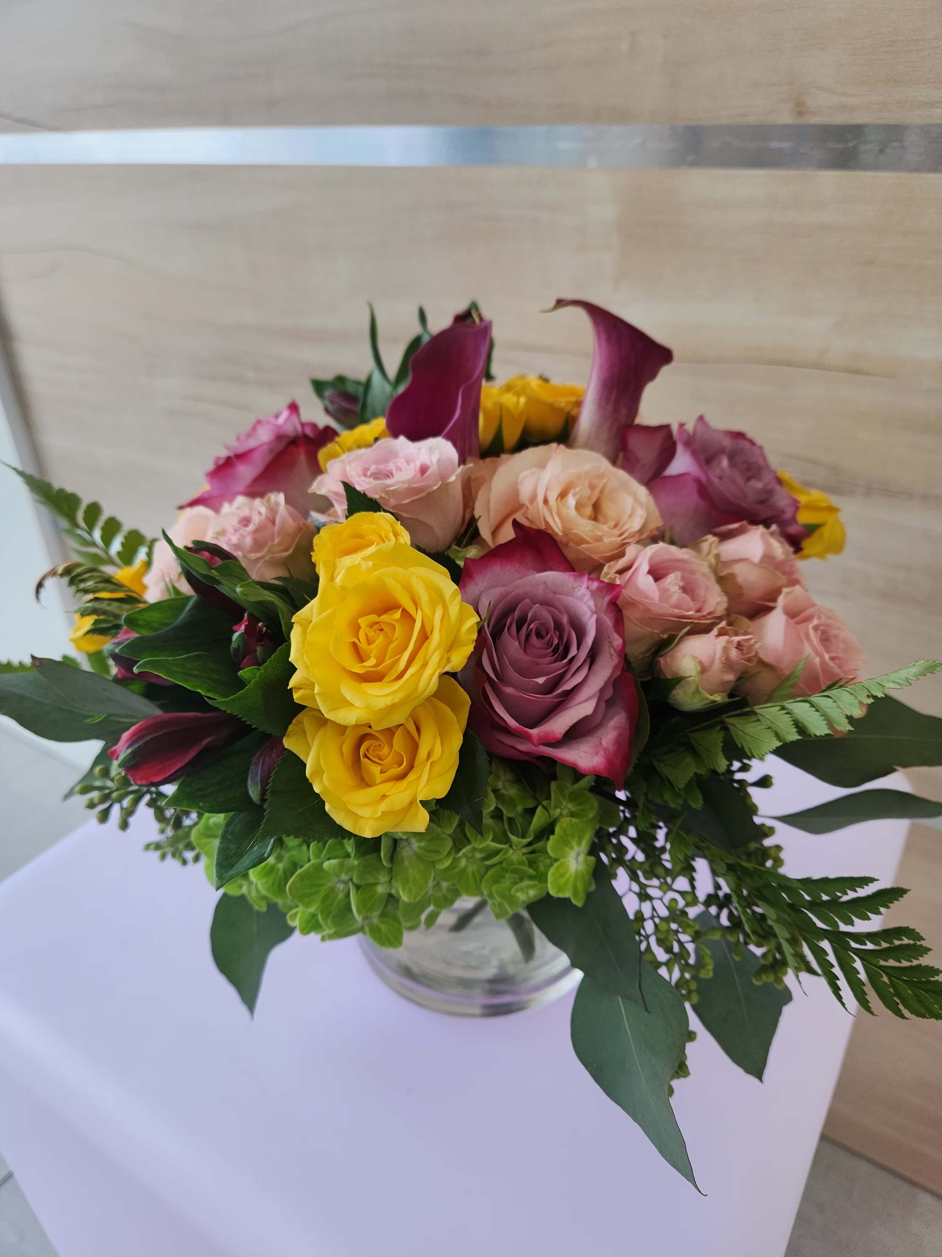 Round arrangement of yellow and pink roses with purple calla lilies in a clear glass vase