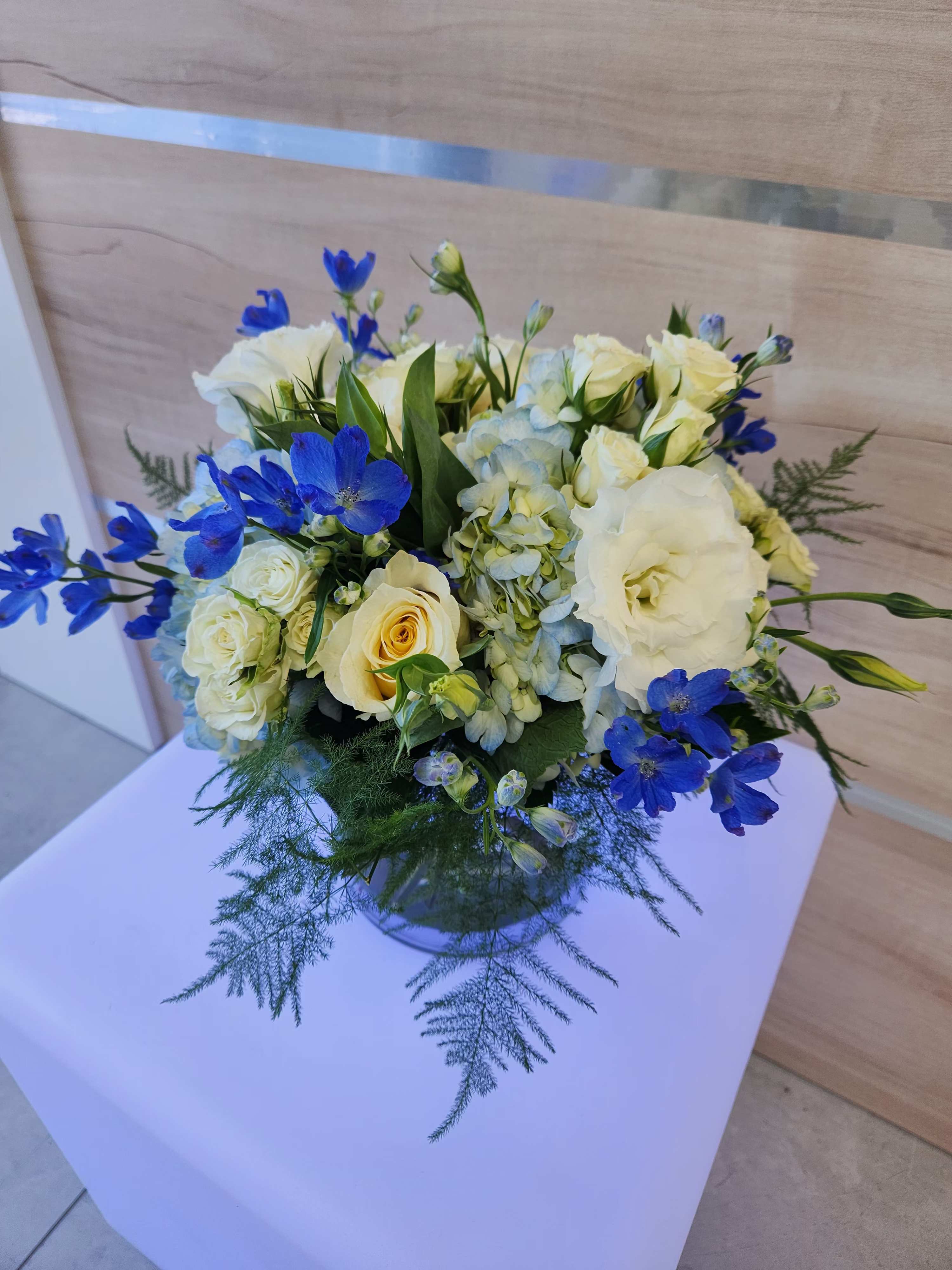 Low round arrangement of white and cream flowers with blue accents in a clear glass vase