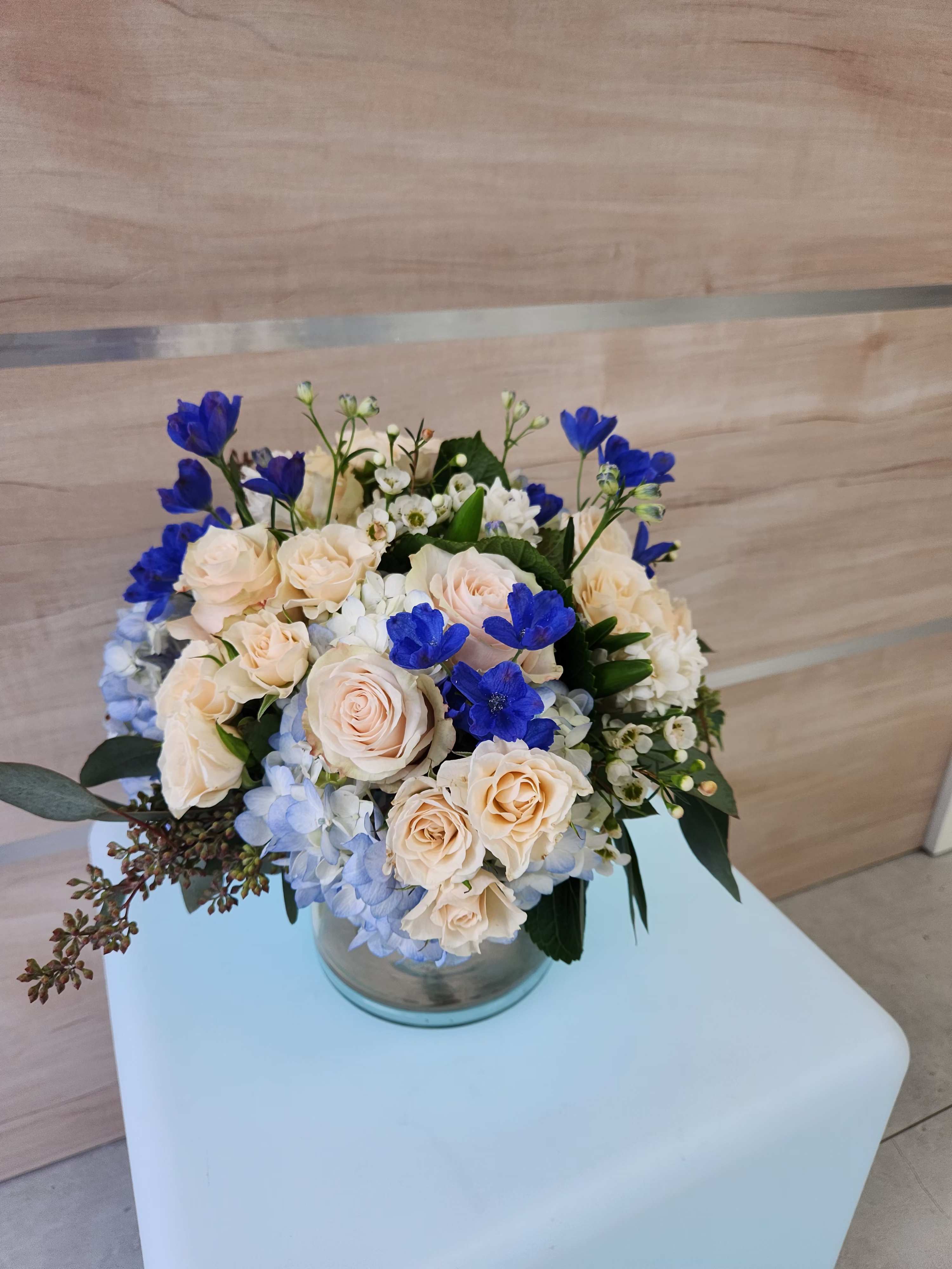 Low bouquet of peach roses, blue hydrangeas, and bright blue flowers in a glass cylinder vase