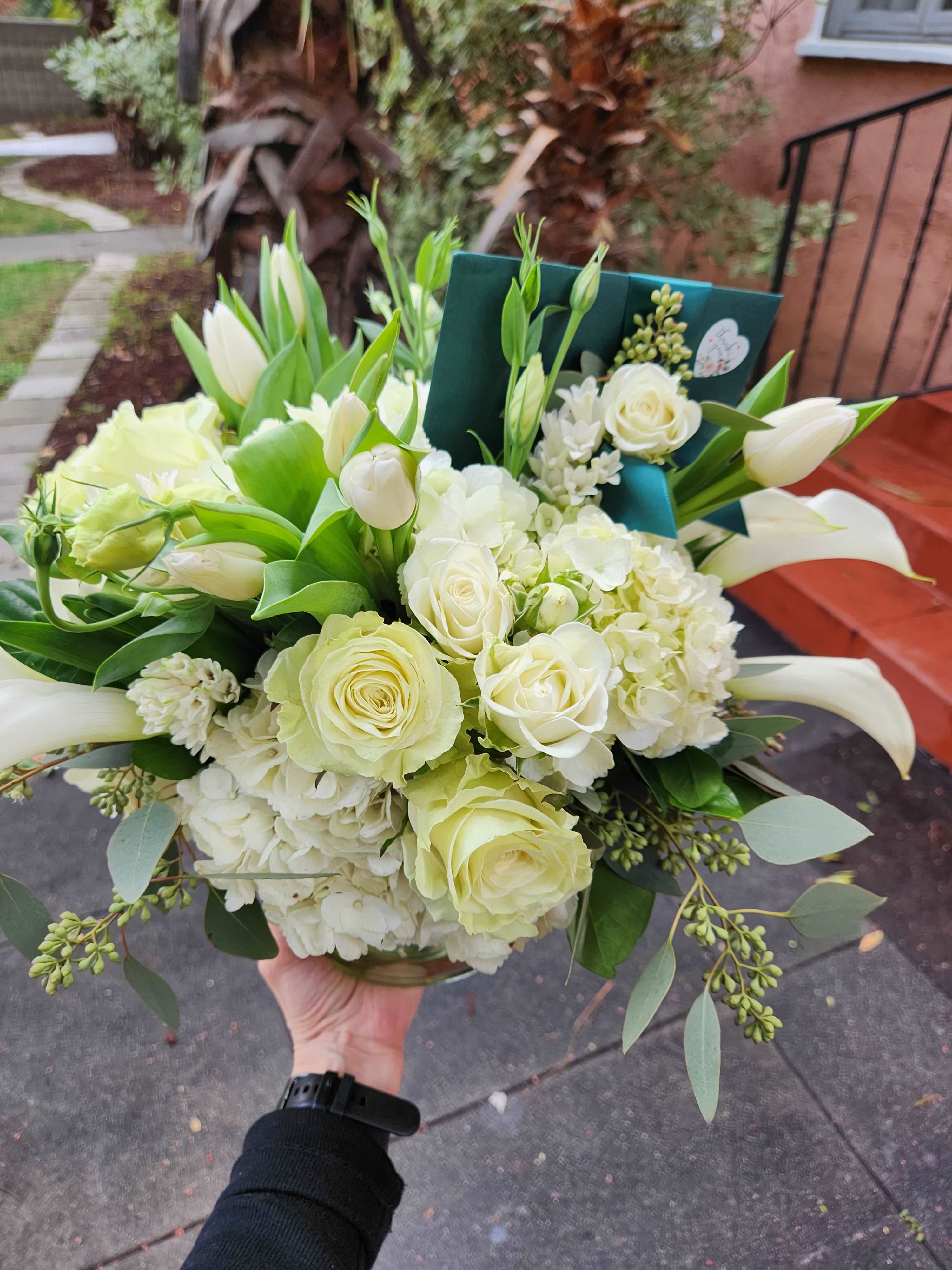 Low arrangement of white roses, hydrangeas, tulips, and calla lilies in a clear glass vase with a green card.