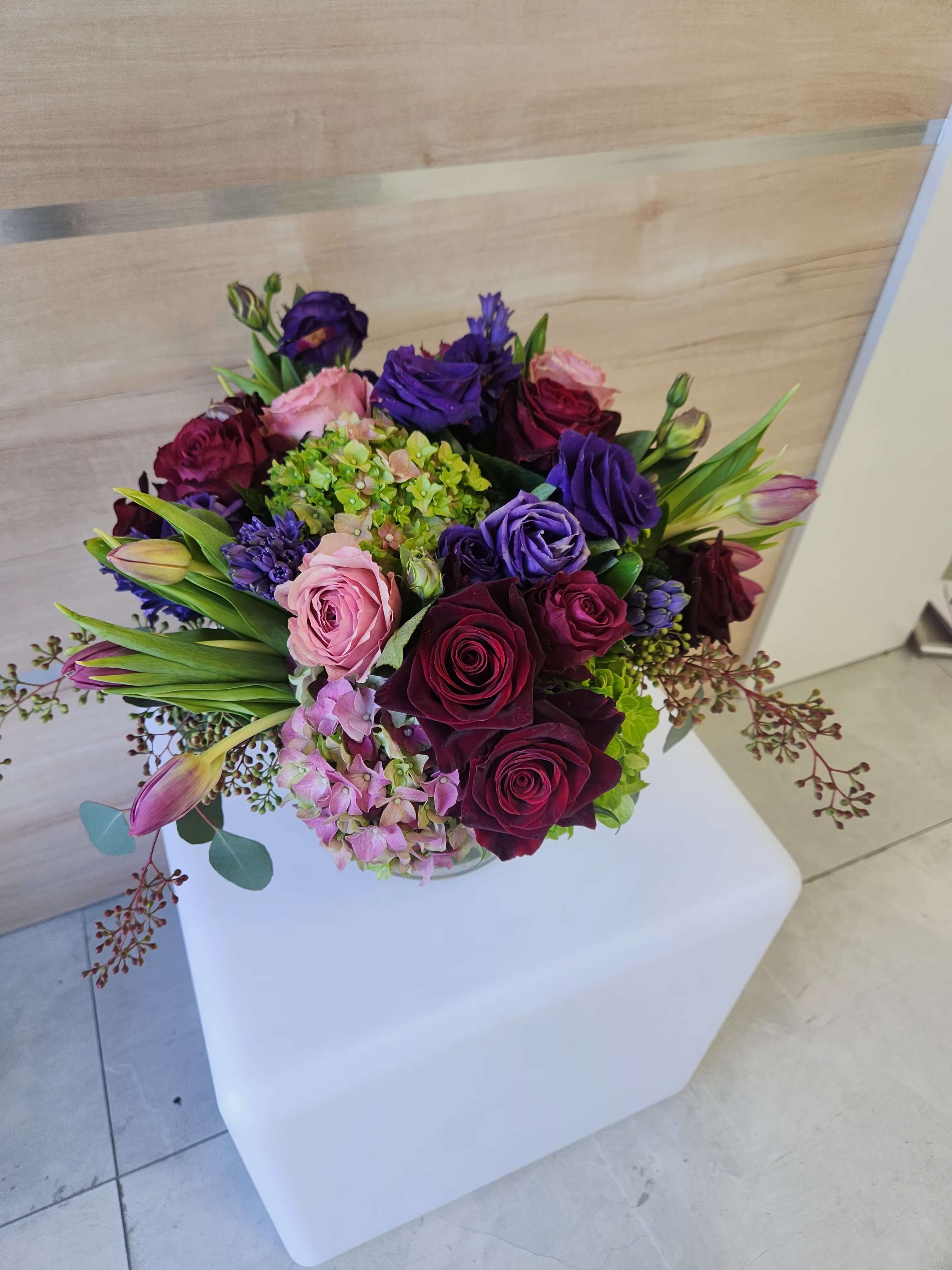 Low round arrangement of red roses, purple lisianthus, tulips and hydrangeas in a clear glass vase