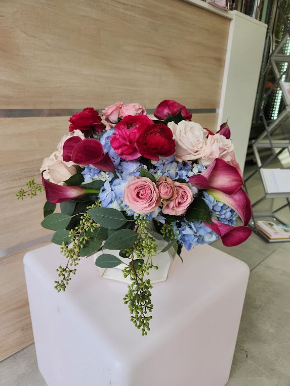 Low arrangement of pink roses, red ranunculus, blue hydrangeas, and dark pink calla lilies in a white vase