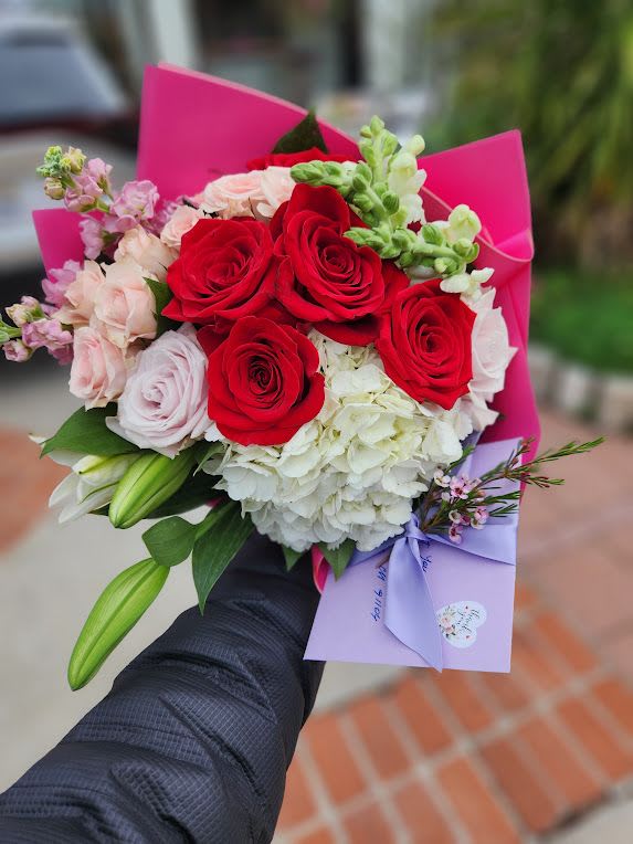 Handheld bouquet of red roses, pink blooms, white hydrangea and lilies with a lavender envelope and pink wrap