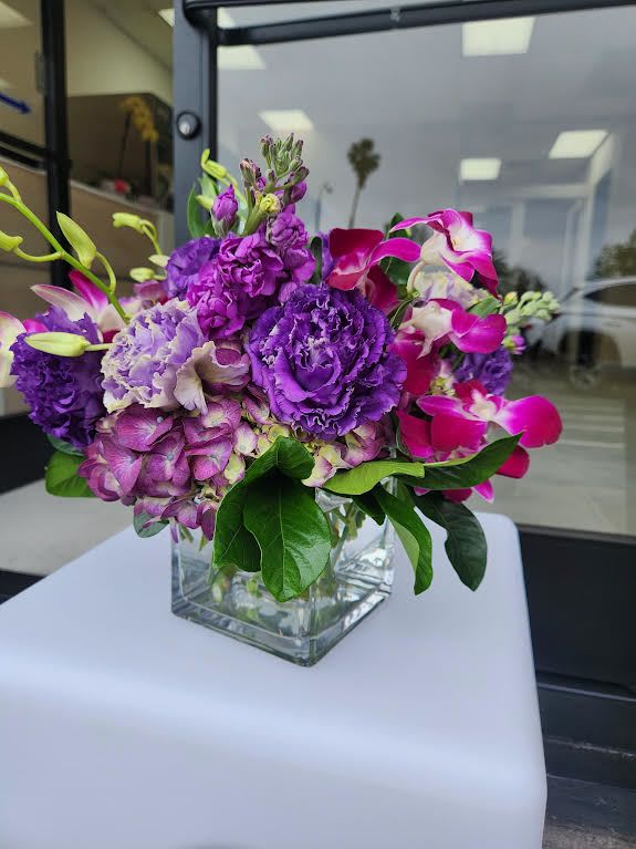 Compact arrangement of purple and pink flowers in a clear glass cube vase.