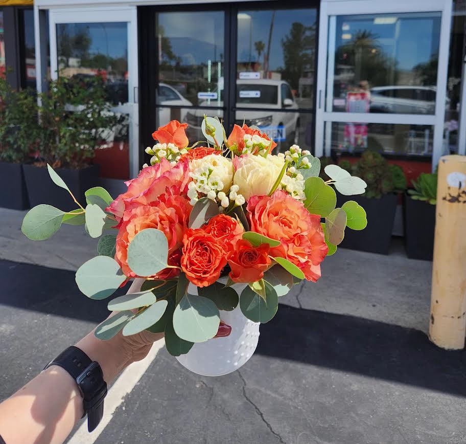 Compact arrangement of orange and cream roses in a small white vase held outdoors