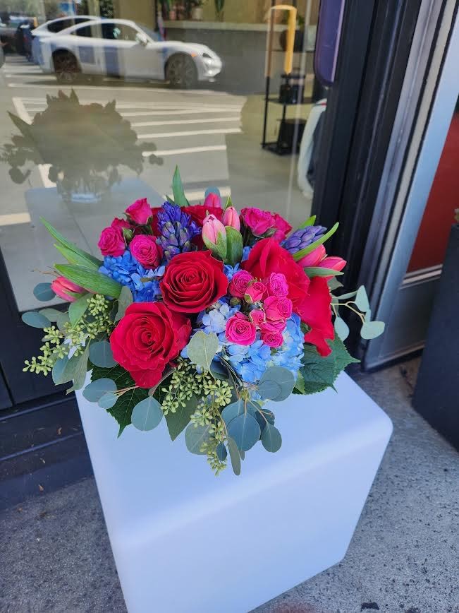 Round arrangement of red roses, pink blooms, blue hydrangea and purple flowers in a low vase
