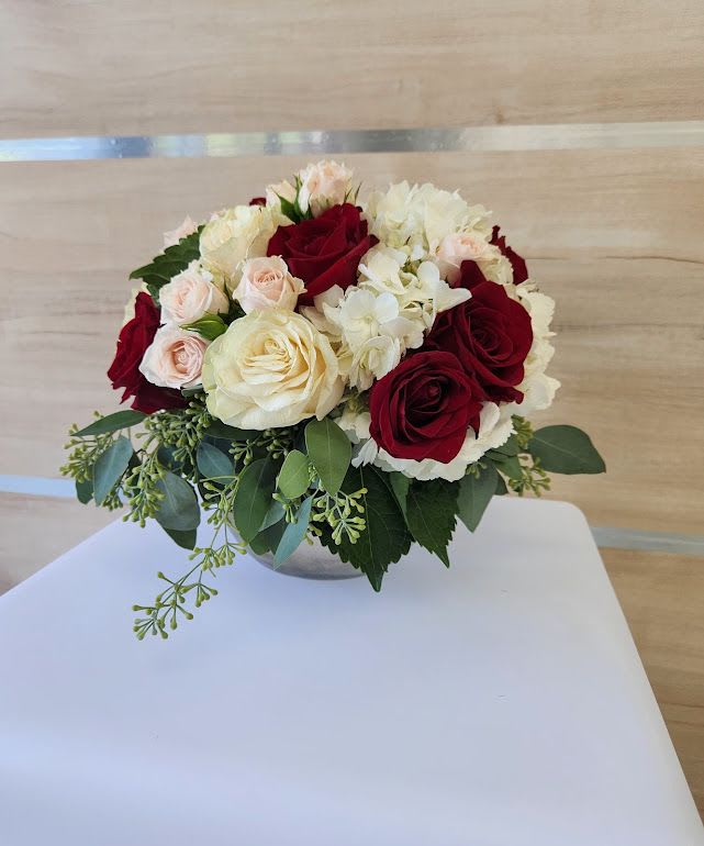 Low round arrangement of red and cream roses, blush spray roses, and white hydrangeas in a small bowl vase.