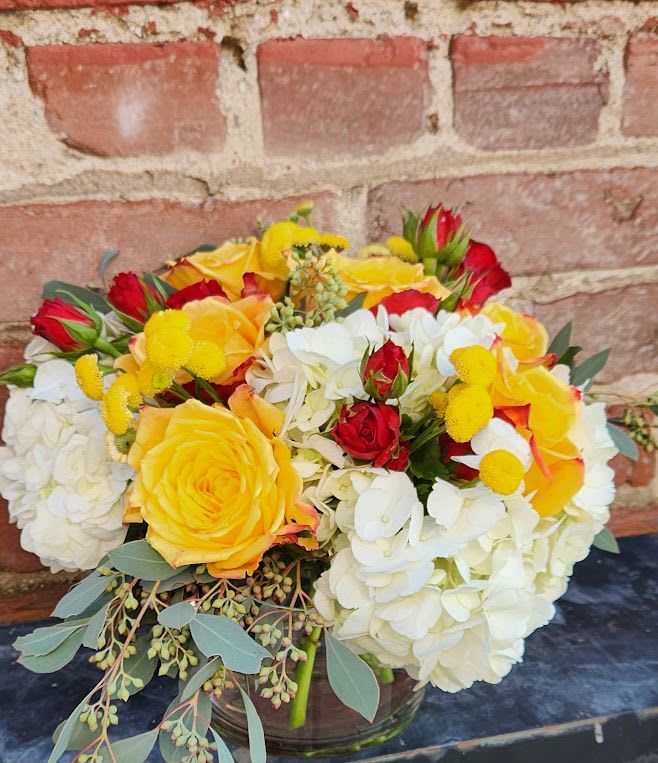Low arrangement of white hydrangeas with yellow and red roses in a clear glass vase