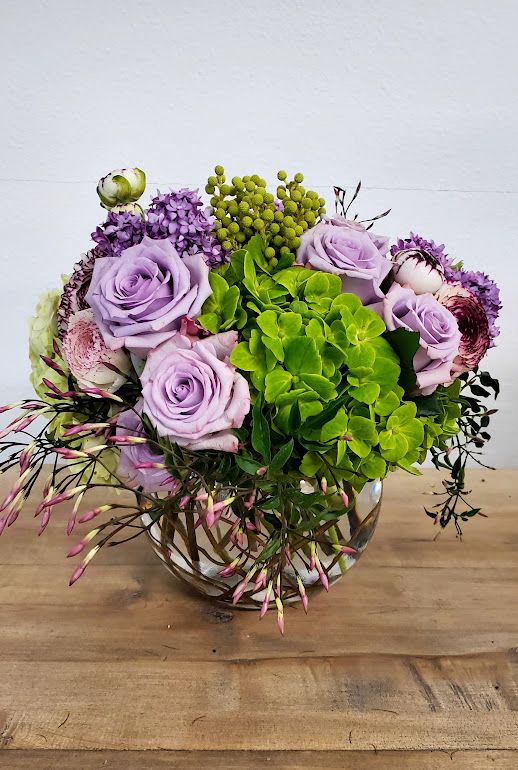 Round arrangement of lavender roses, green hydrangea, and mixed blooms in a clear glass bowl vase