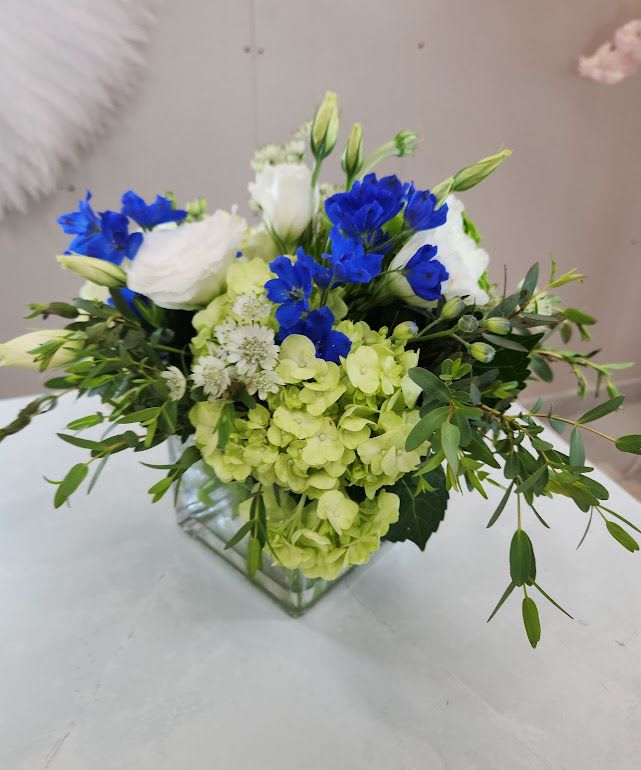 Low arrangement of green hydrangeas, blue delphinium, and white blooms in a clear glass cube vase